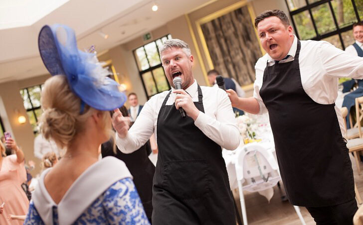 Two singing waiters performing to a guest in a hat at a wedding
